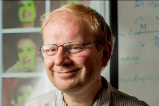 Alexei A. Efros mit Brille lächelt, während er vor einer Tafel mit Gesichtserkennungsbildern und Schriftzeichen steht.