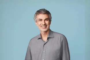 Yoshua Bengio mit grauen Haaren und Bart, bekleidet mit einem grauen Hemd mit Knöpfen, vor einem hellblauen Hintergrund.