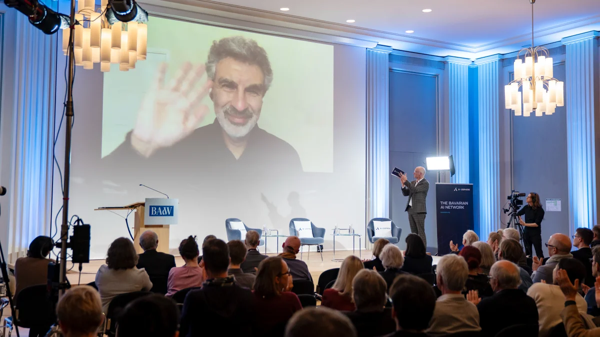 A man waves on a large screen during a conference in a well-lit auditorium, with an audience and a speaker on stage.