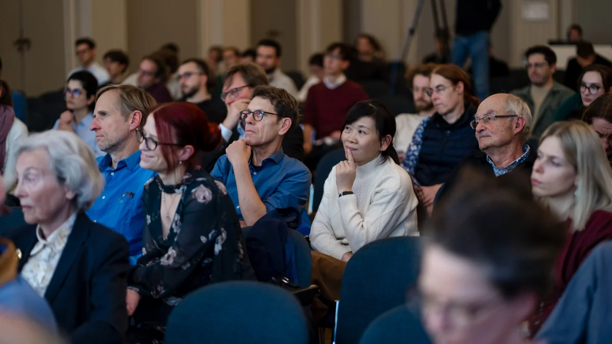 Zuschauer sitzen und hören während einer Veranstaltung in einem Innenraum aufmerksam zu, mit unterschiedlichen Gesichtsausdrücken und legerer Kleidung.