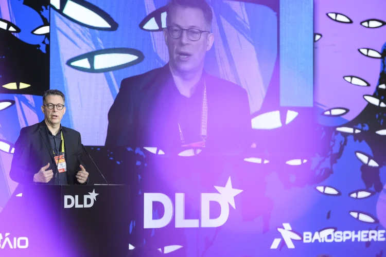 Markus Blume speaks on stage at a conference with "DLD" branding, colorful lights, and eye-shaped patterns in the background.