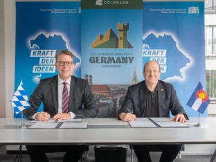 Two men in suits sit at a table with signed documents, flanked by Bavarian and Colorado flags, in front of banners for a Germany 2026 mission.