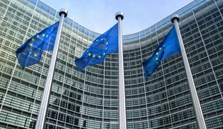 Three European Union flags on poles in front of a modern glass building with a curved facade under a blue sky.