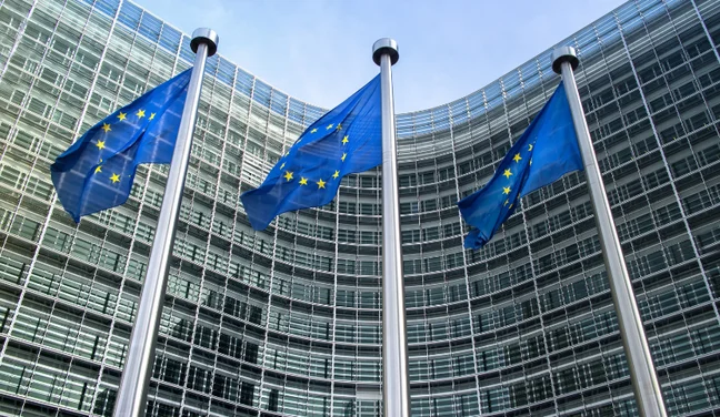 Three European Union flags on poles in front of a modern glass building with a curved facade under a blue sky.