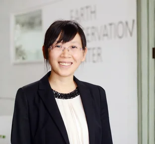 Xiaoxiang Zhu mit Brille und schwarzem Blazer steht vor einem unscharfen Hintergrund mit Text an einer Wand.