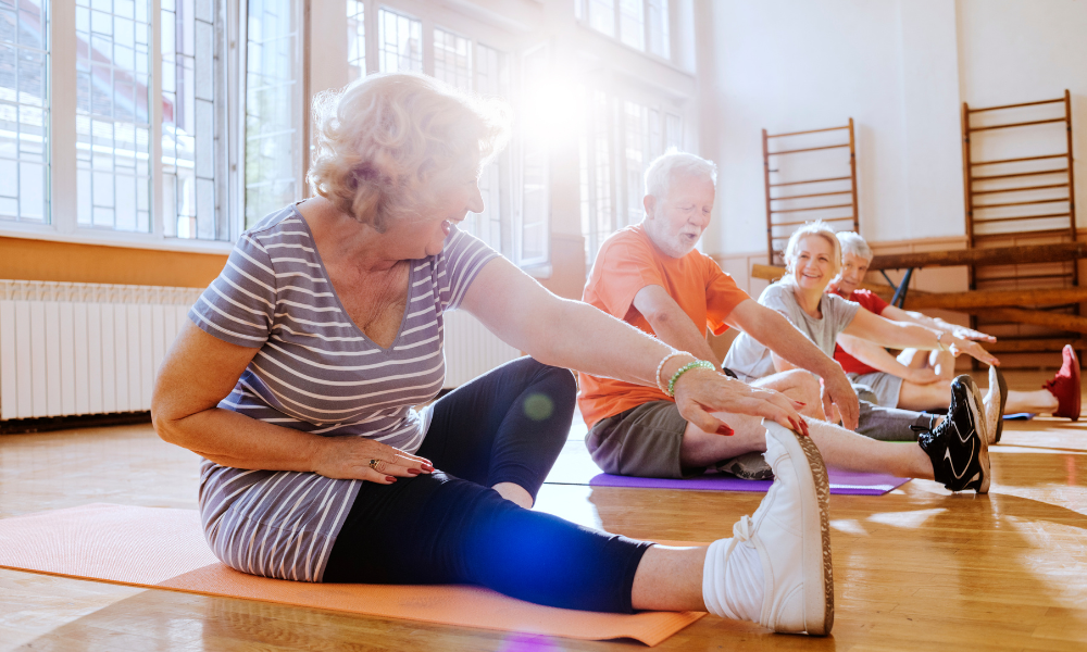 Seniors in a bright room doing seated stretches on yoga mats, smiling and reaching for their toes in a sunlit exercise space.