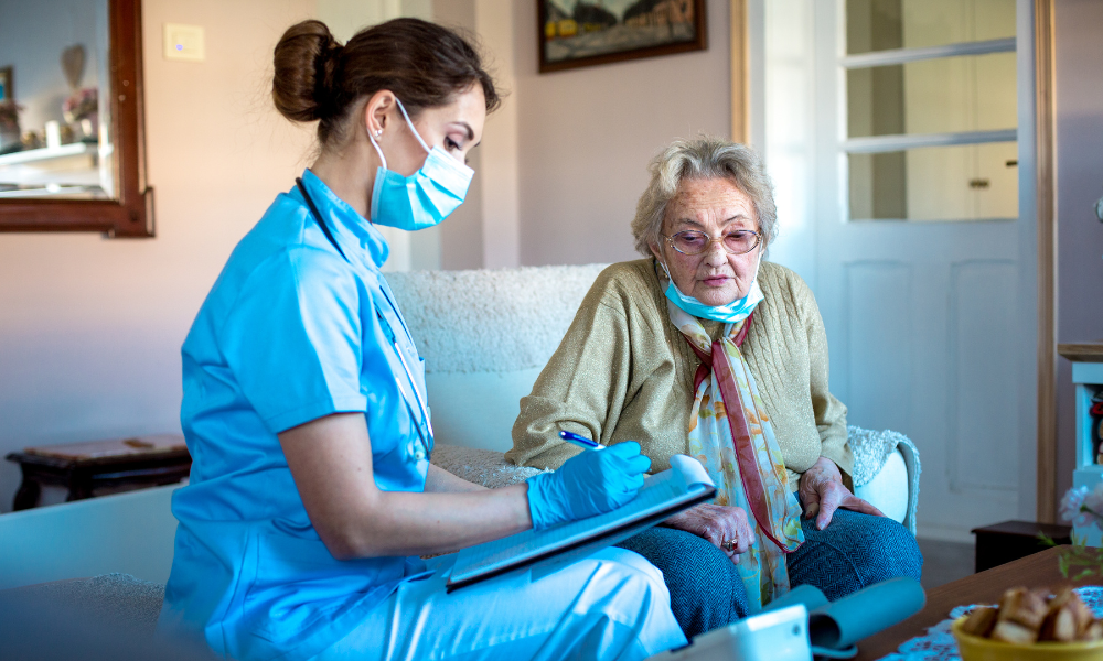 A nurse in blue scrubs and mask writes on a clipboard while sitting beside an elderly woman wearing glasses and a mask, in a cozy room.