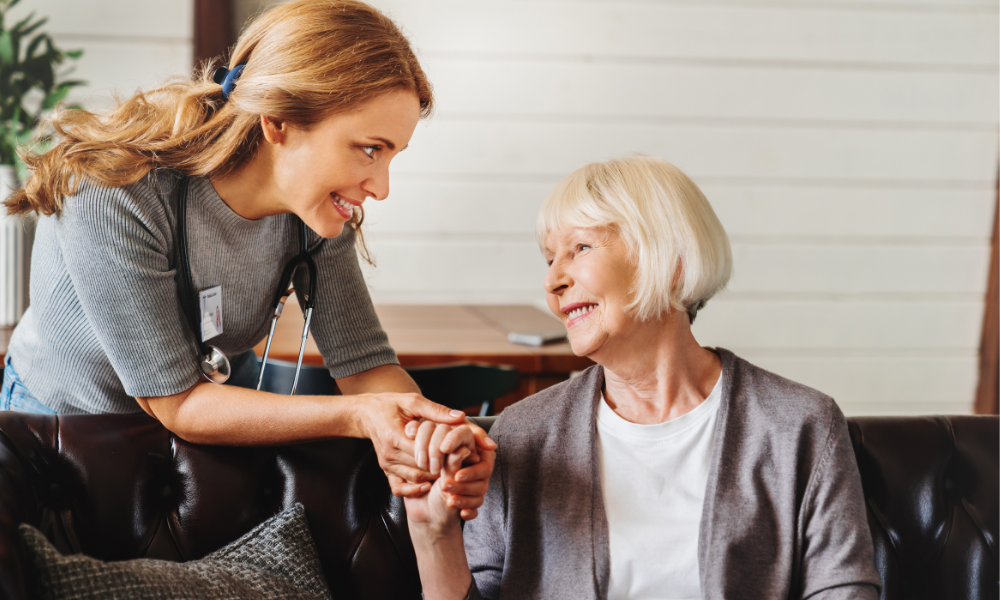 A smiling caregiver with a stethoscope holds hands with an elderly woman, sharing a warm moment on a couch in a cozy room.