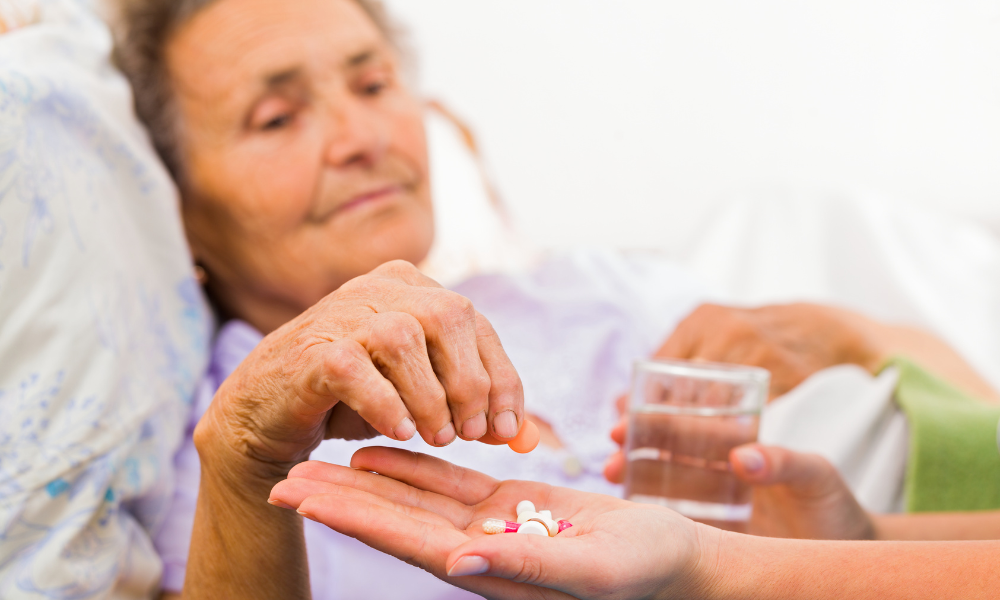 Elderly woman in bed takes pills from a caregiver's hand, holding a glass of water.