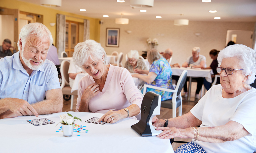 Elderly people playing bingo at a table in a well-lit room, with others engaged in similar activities in the background.