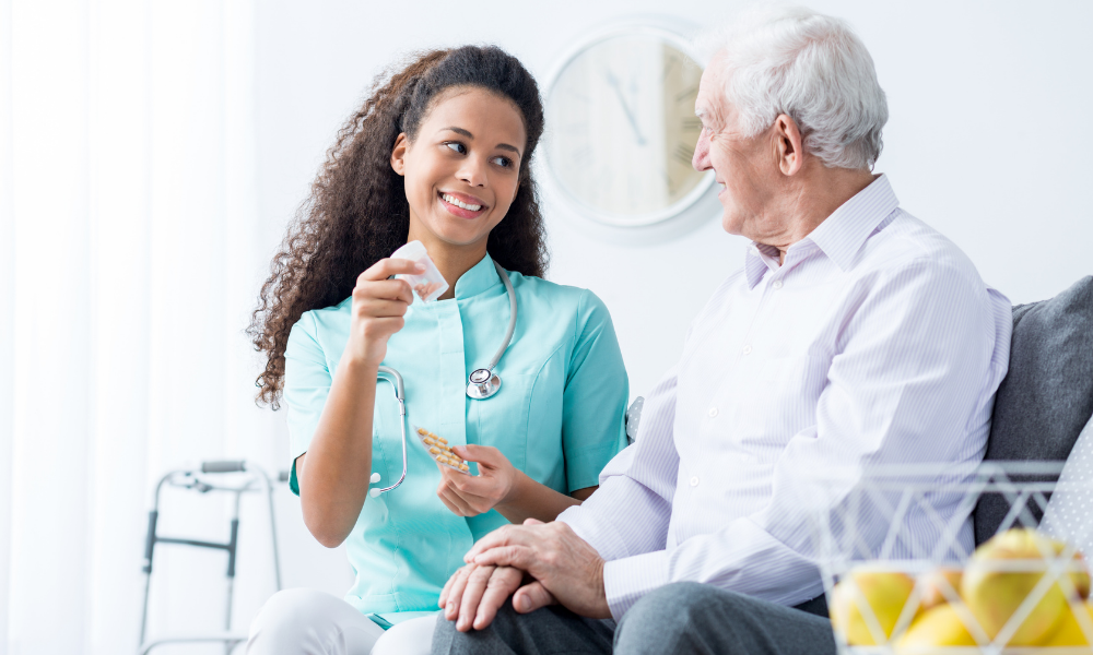 A nurse with a stethoscope smiles at an elderly man while holding medication. They sit in a bright room with a walker and fruit basket nearby.