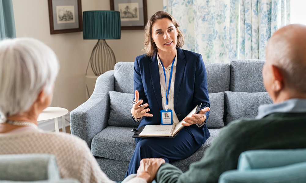 A professional woman in a suit talks to an elderly couple sitting on a sofa, holding hands, in a cozy living room setting.