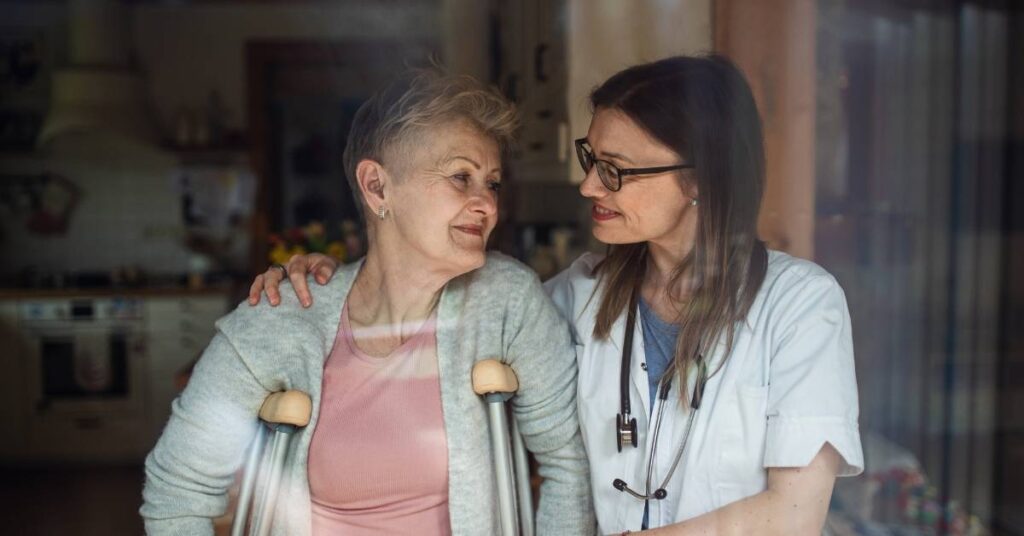 A healthcare professional comforts an elderly woman using crutches, both smiling at each other warmly in a home setting.