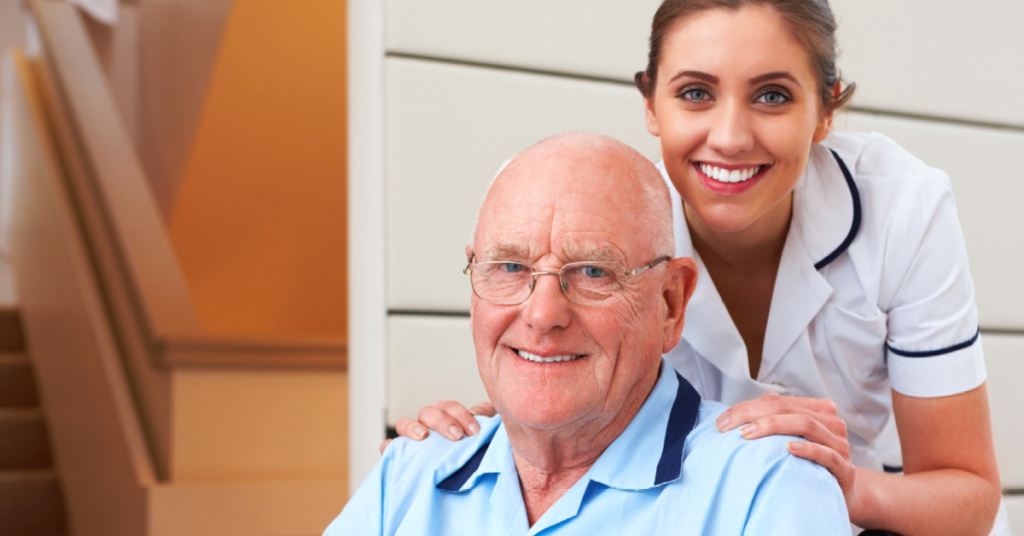 Elderly man smiling with a young caregiver in a white uniform, both looking at the camera indoors.