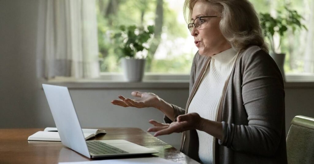 Older woman with glasses gestures at a laptop while sitting at a table, looking confused. Bright room with plants in the background.