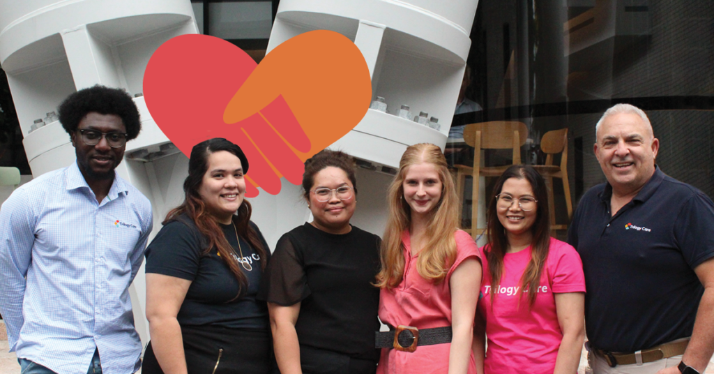 A diverse group of six people smiling, standing in front of a building with a large heart graphic in the background.