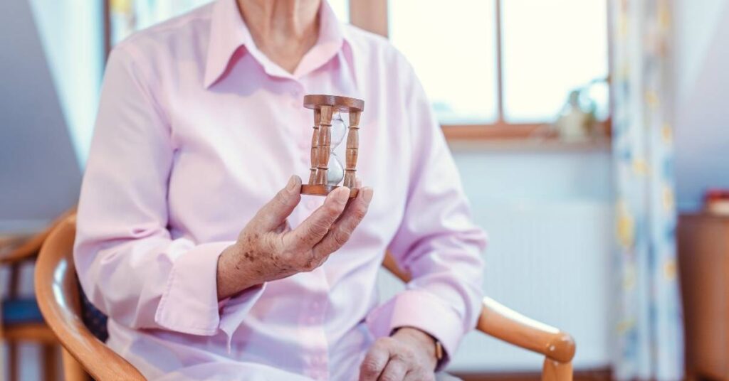 Elderly person in a pink shirt holding a small wooden hourglass while sitting on a chair indoors.