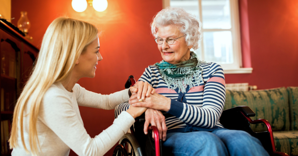 A young woman kneels, holding hands with an elderly woman in a wheelchair, both smiling in a warmly lit room.