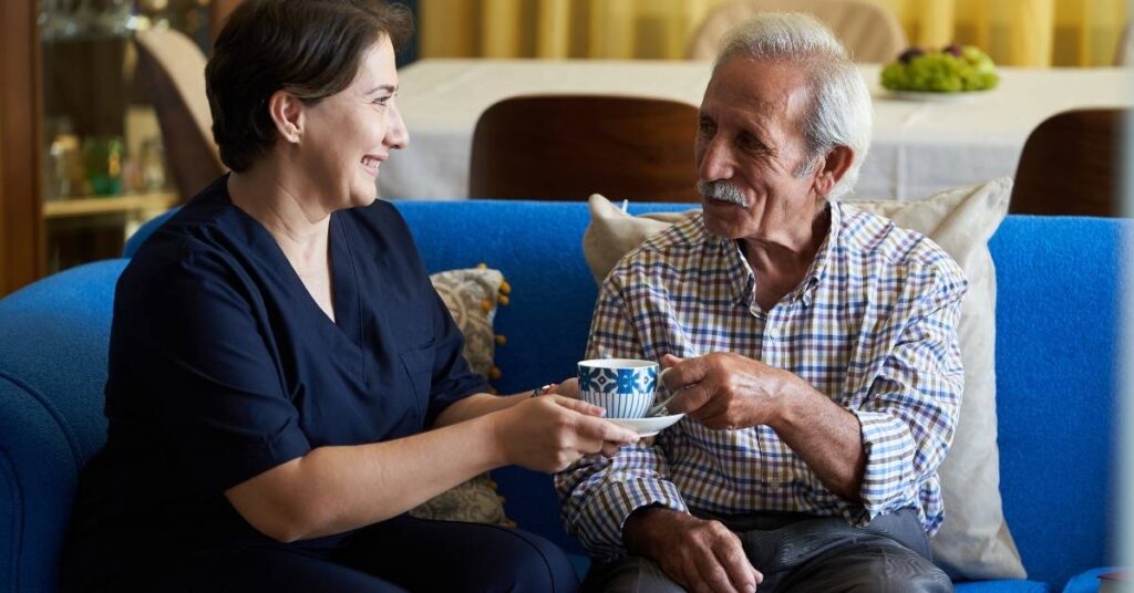A smiling woman hands a cup to an elderly man on a blue sofa, with a dining table in the background.