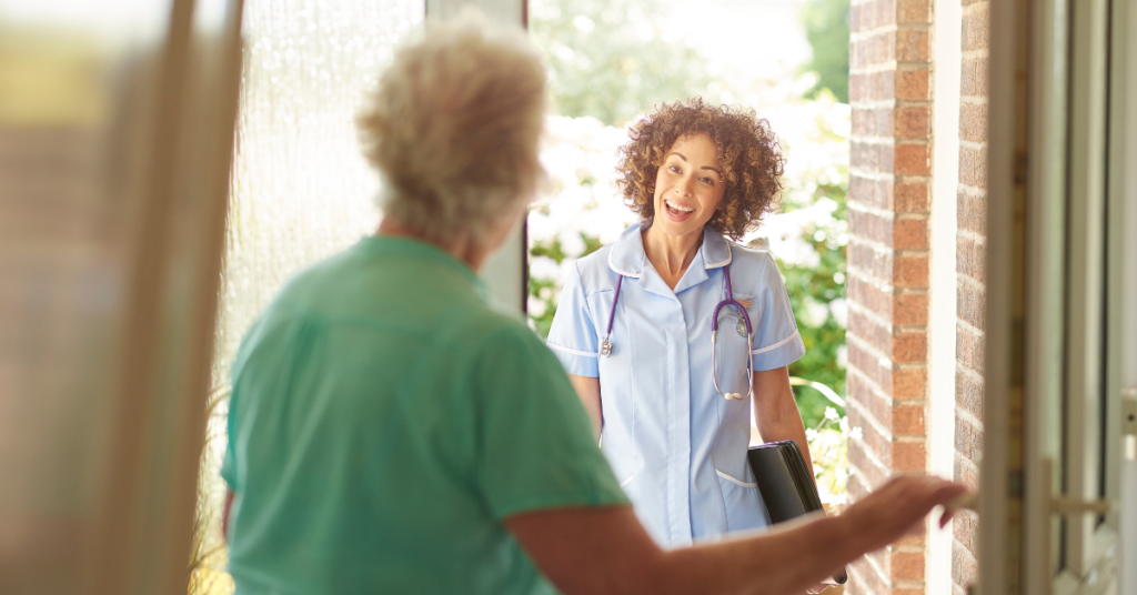A nurse with a stethoscope smiles at an elderly person at the door, holding a folder. Sunlight filters through trees in the background.