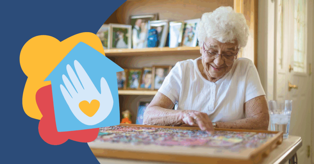 Elderly woman with white hair working on a colorful puzzle at a table, surrounded by photographs. A glass of water is nearby.