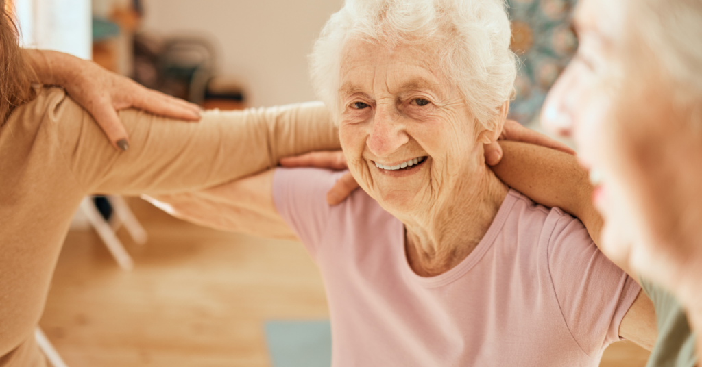 Elderly woman smiling during a group exercise class, arms linked with others, wearing a light pink shirt in a bright room.
