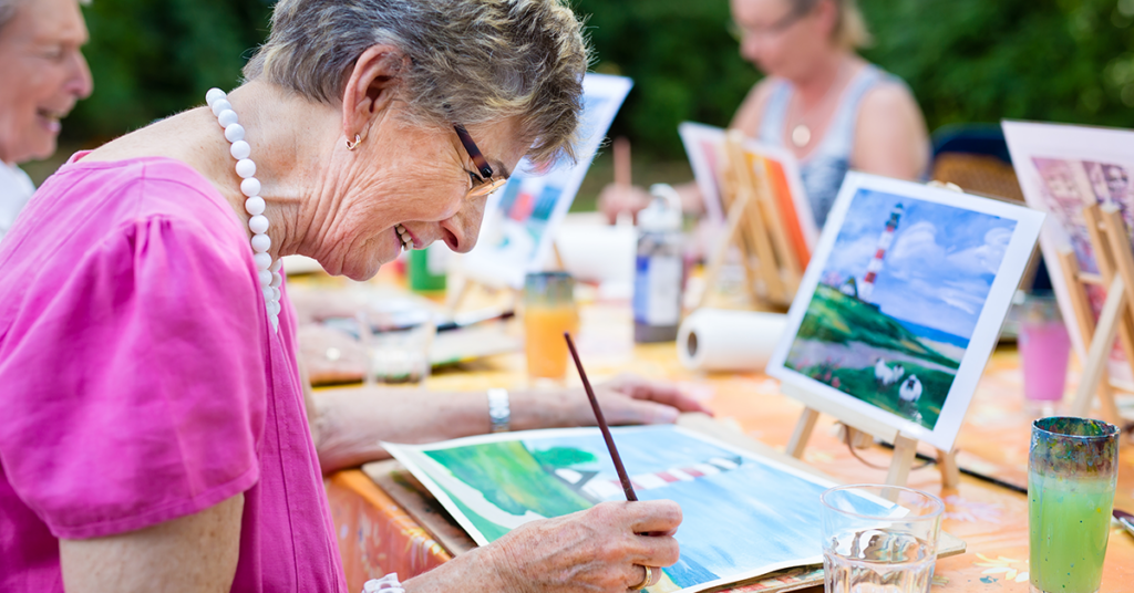 Elderly woman in a pink blouse painting a landscape on canvas outdoors, surrounded by other artists and art supplies.
