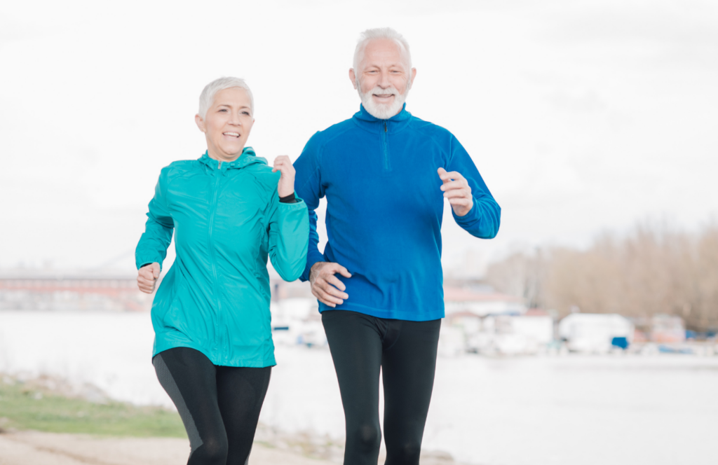 Older couple jogging outdoors near a river, wearing blue jackets and black pants, with a blurred background of trees and buildings.