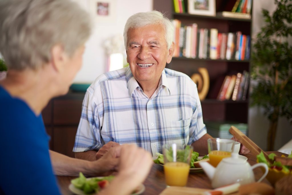 Elderly couple enjoying a meal together at a table with salads, juice, and a teapot, in a cozy room with bookshelves in the background.