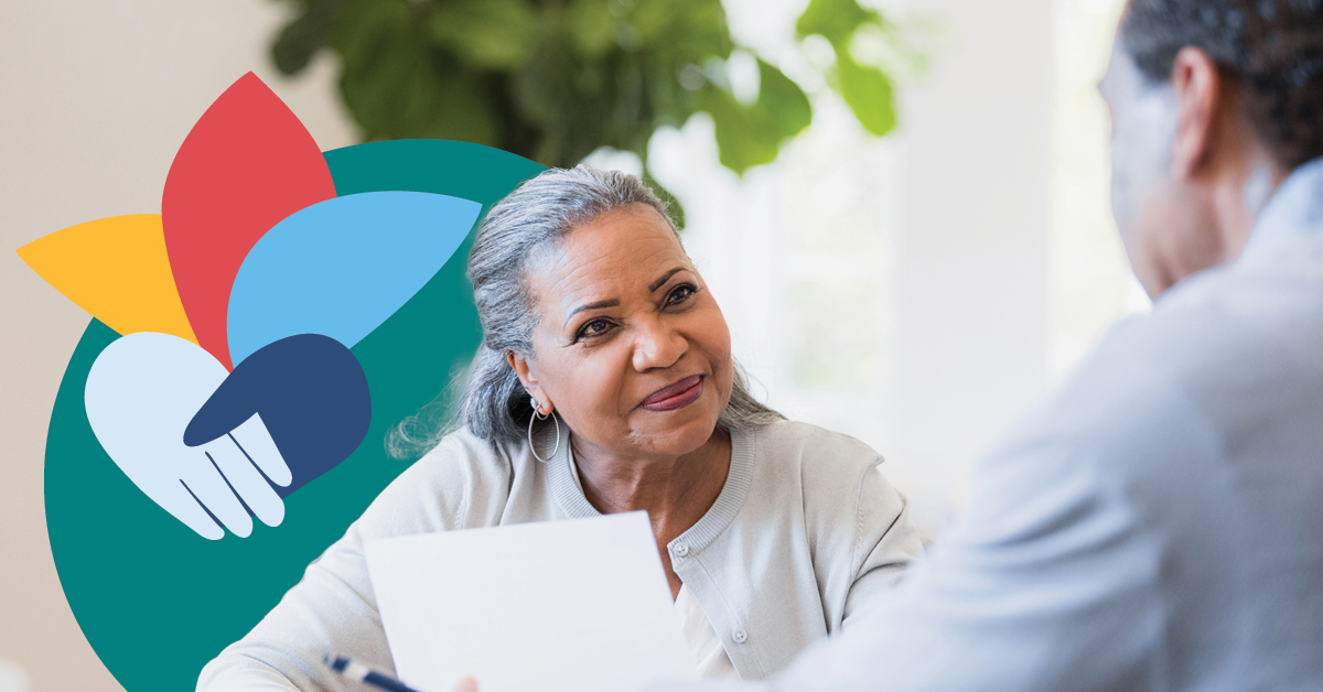 Older woman holding a paper, smiling at a man. Colorful flower and handshake logo in the background. Bright, indoor setting.