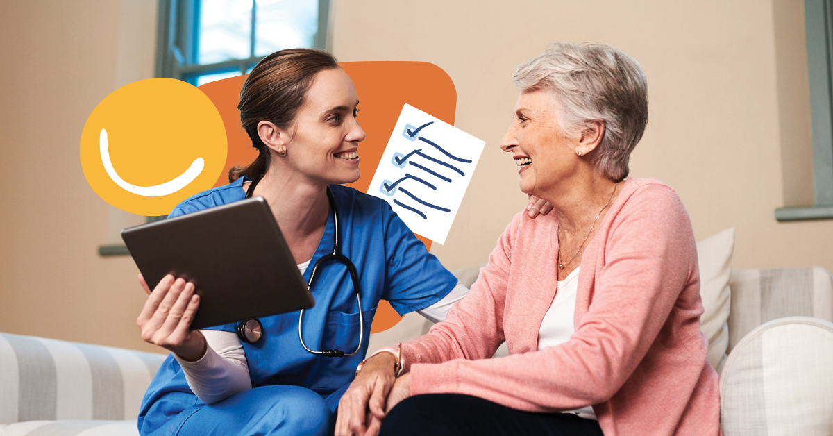 Nurse in blue scrubs holding a tablet, smiling at an elderly woman in a pink cardigan, sitting on a couch in a cozy room.