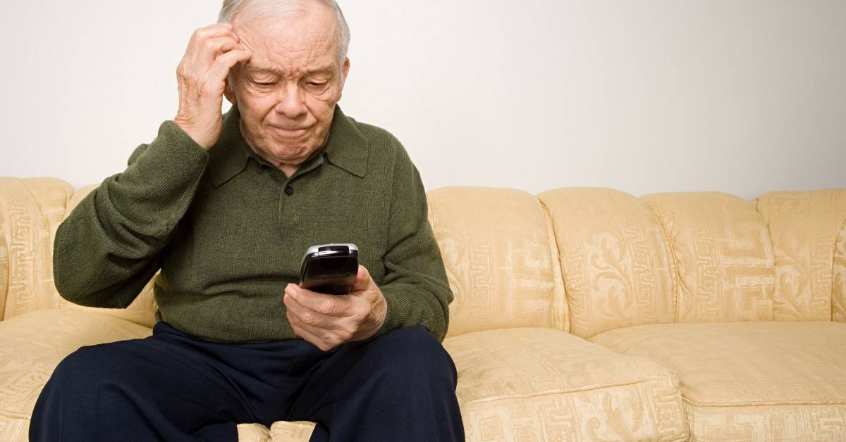 Elderly man sitting on a beige sofa, looking confused while holding a smartphone, scratching his head.