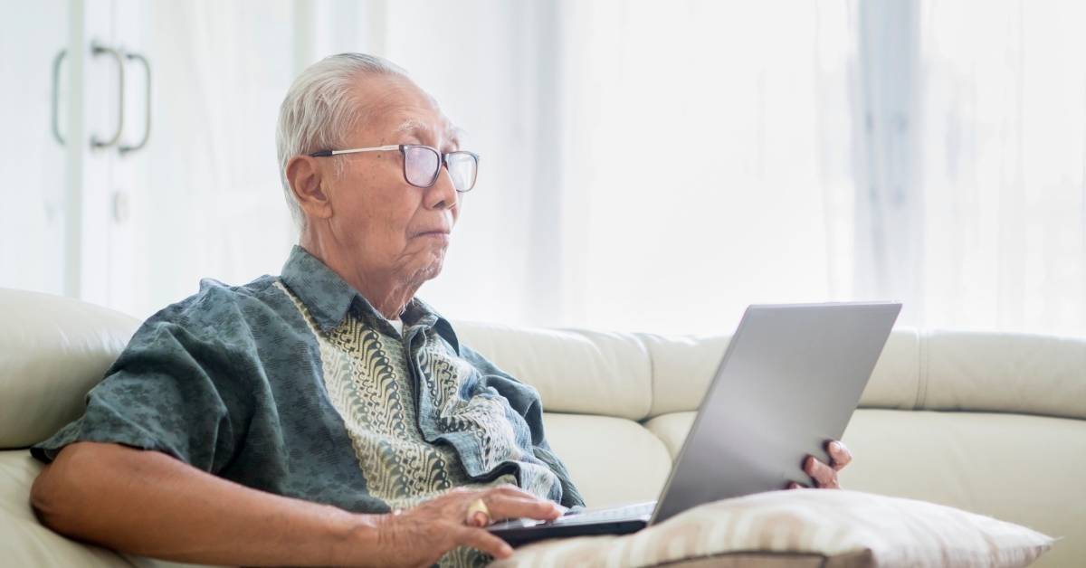 Elderly person with glasses sitting on a sofa, using a laptop, in a bright room with light curtains.