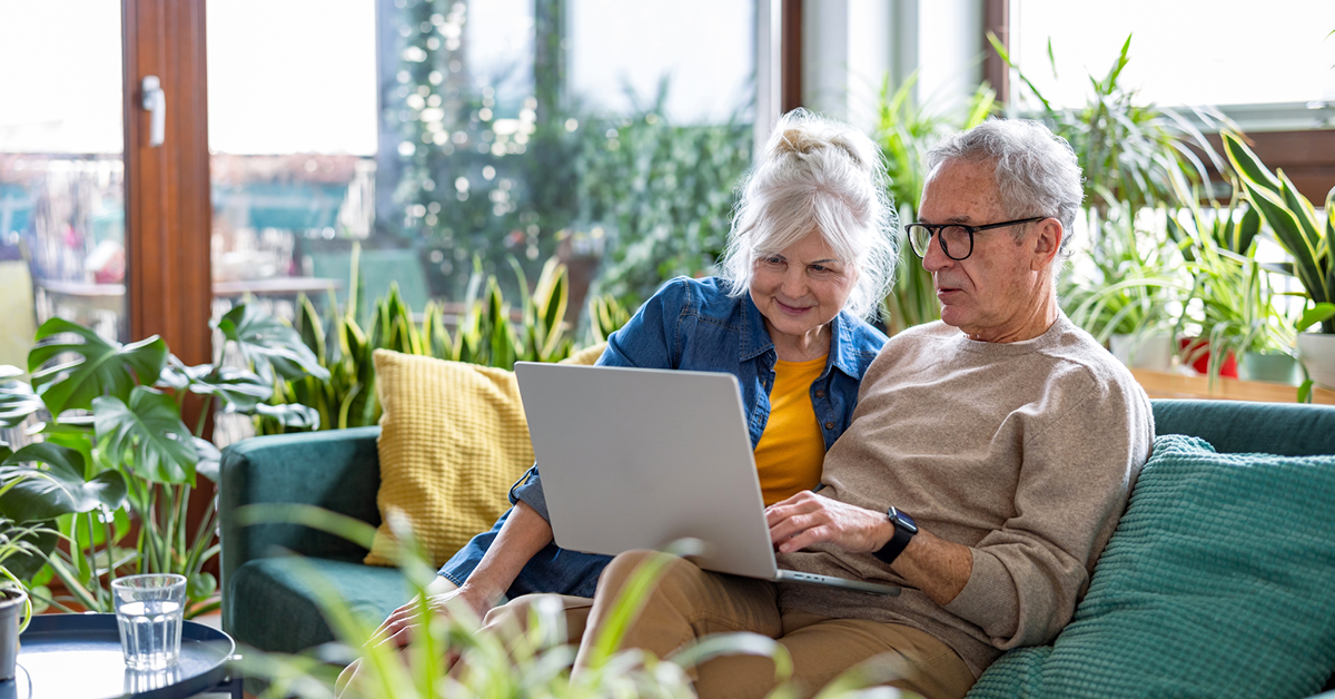 Elderly couple sitting on a sofa surrounded by plants, using a laptop together, appearing engaged and content.