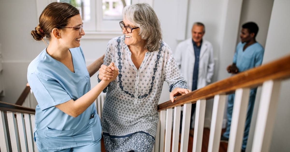 A nurse assists an elderly woman up the stairs, while two healthcare professionals follow behind in a well-lit hallway.