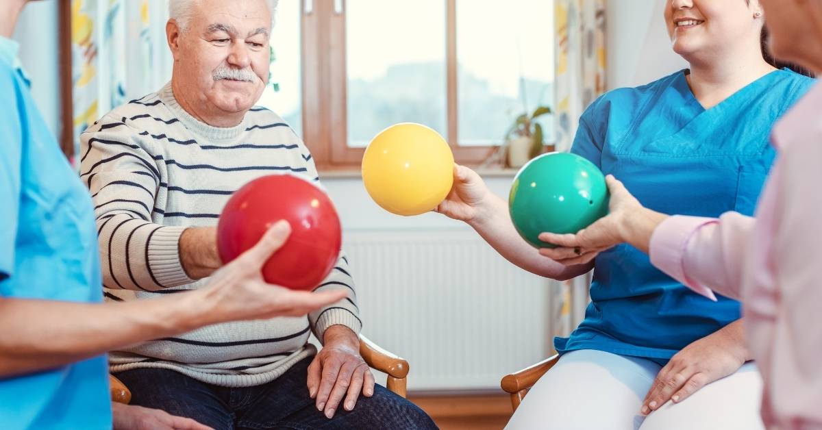 Elderly man and caregivers in a room passing colorful balls during a group activity, promoting physical engagement and social interaction.
