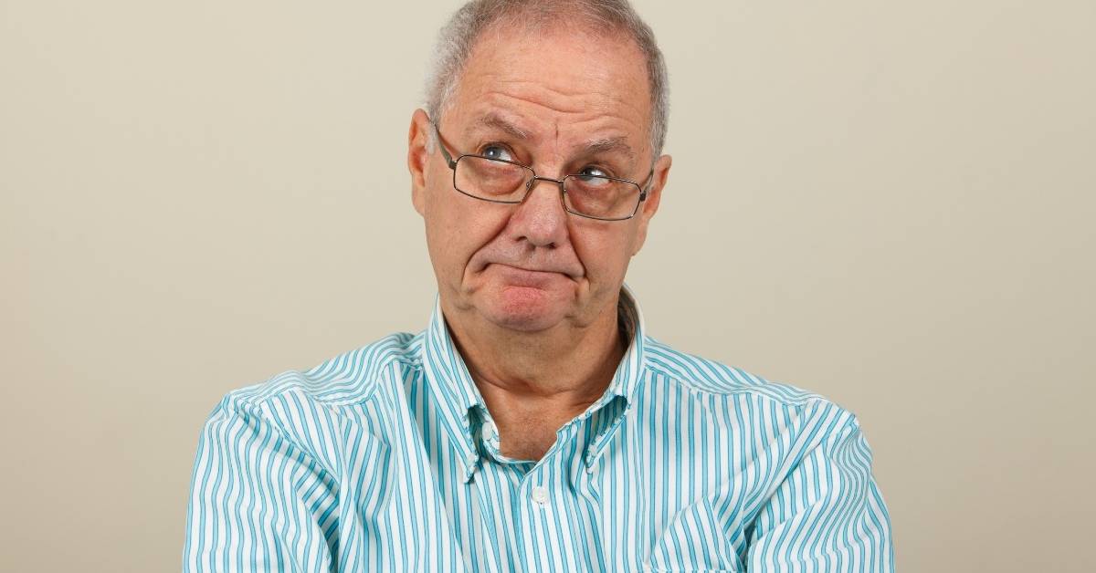 Elderly man with glasses and a striped shirt looks up with a puzzled expression against a plain background.
