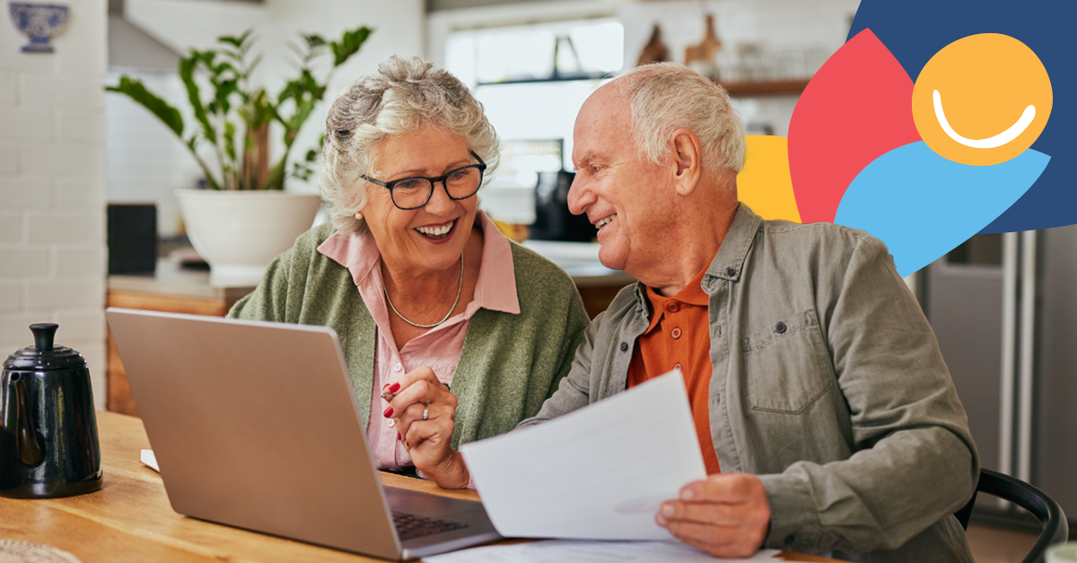 Elderly couple smiling while using a laptop at a kitchen table, holding papers. Colorful abstract shapes in the background.