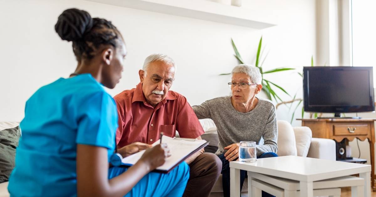 Healthcare worker in blue scrubs discusses paperwork with an older couple sitting on a couch in a living room.