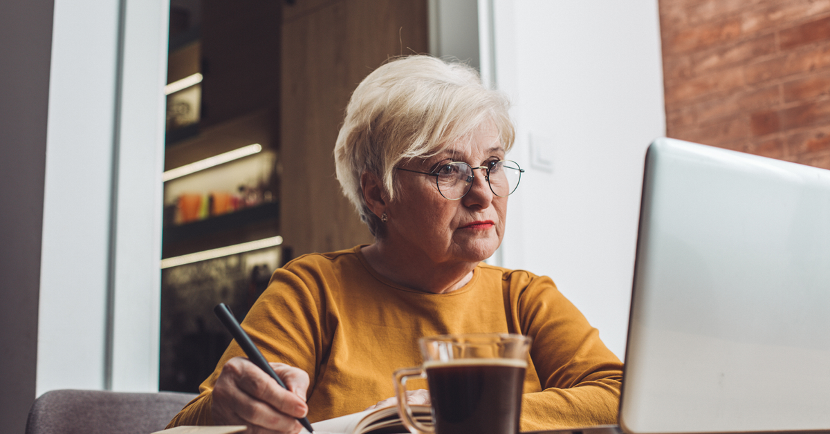 Elderly woman with glasses writing in a notebook while looking at a laptop, with a cup of coffee on the table in a cozy room.