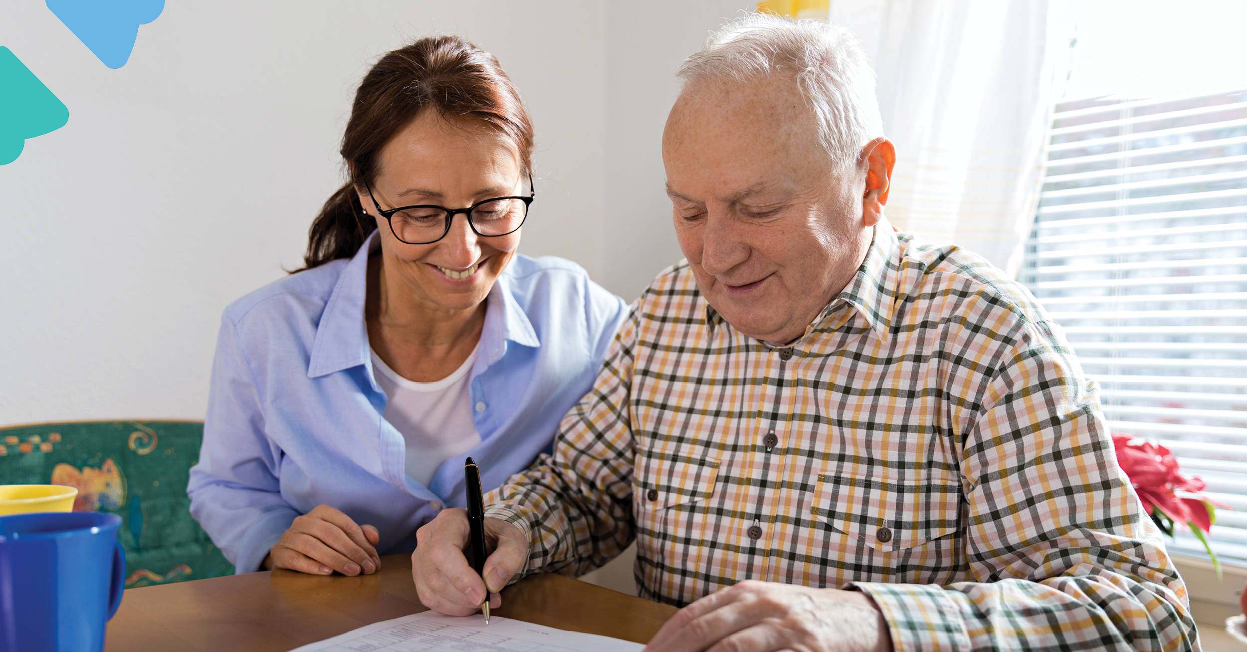 Elderly man in a plaid shirt signing a document with a smiling woman in glasses assisting him at a table.