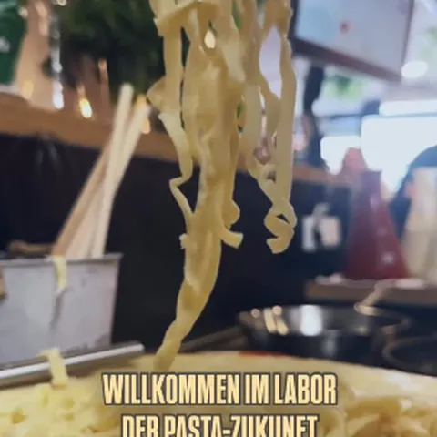 Frische Nudeln werden in einer Küche mit einer Zange herausgehoben. Der deutsche Text lautet: „Willkommen im Labor der Pasta-Zukunft“.