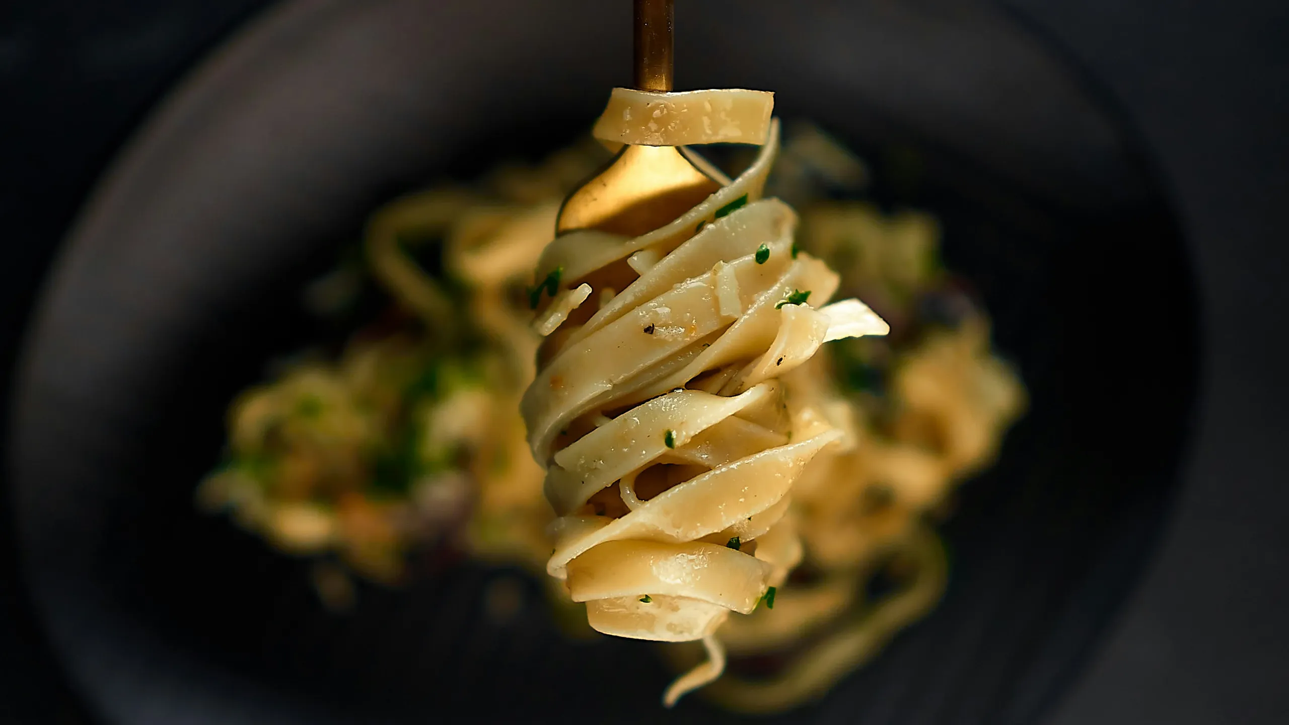 Eine Gabel wirbelt cremige, mit Kräutern garnierte Fettuccine-Nudeln über einem dunklen Teller, auf dem im Hintergrund weitere Nudeln liegen.