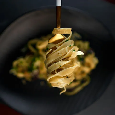 Eine Gabel wirbelt cremige, mit Kräutern garnierte Fettuccine-Nudeln über einem dunklen Teller, auf dem im Hintergrund weitere Nudeln liegen.