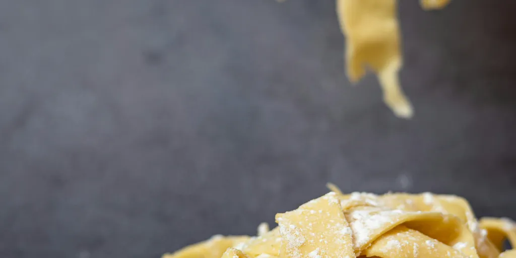 Ein Haufen ungekochter, mit Mehl bestäubter Fettuccine-Nudeln auf einer dunklen Fläche, mit darüber hängenden Fäden.