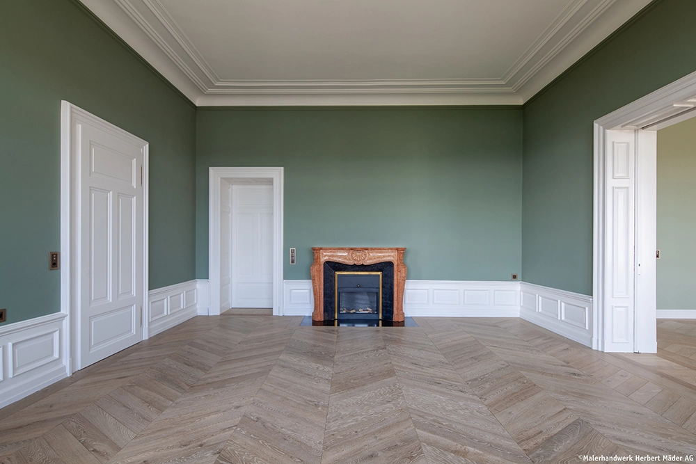 Spacious room with green walls, white trim, herringbone wood floor, and a decorative marble fireplace between two white doors.