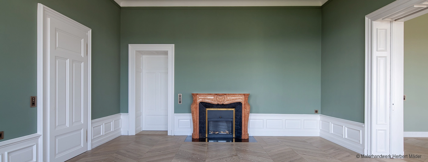 Spacious room with green walls, white trim, herringbone wood floor, and a decorative marble fireplace between two white doors.