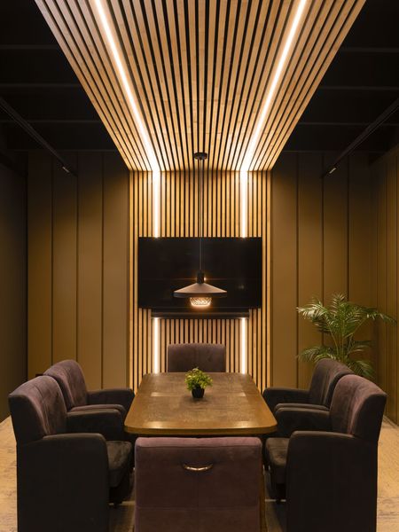 Modern meeting room with a wooden table, plush chairs, vertical slat walls, ceiling lights, a mounted screen, and a small plant centerpiece.