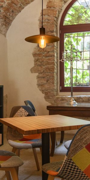 Cozy room with a modern wooden table, patchwork chairs, exposed brick wall, large arched window, and vintage hanging light bulb.