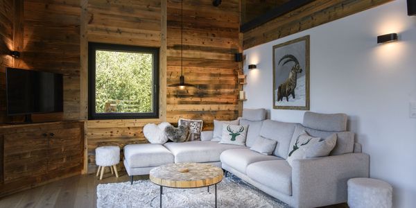 Cozy living room with wooden walls, gray sectional sofa, deer-themed cushions, fluffy rug, TV, and goat artwork. Large window with a nature view.